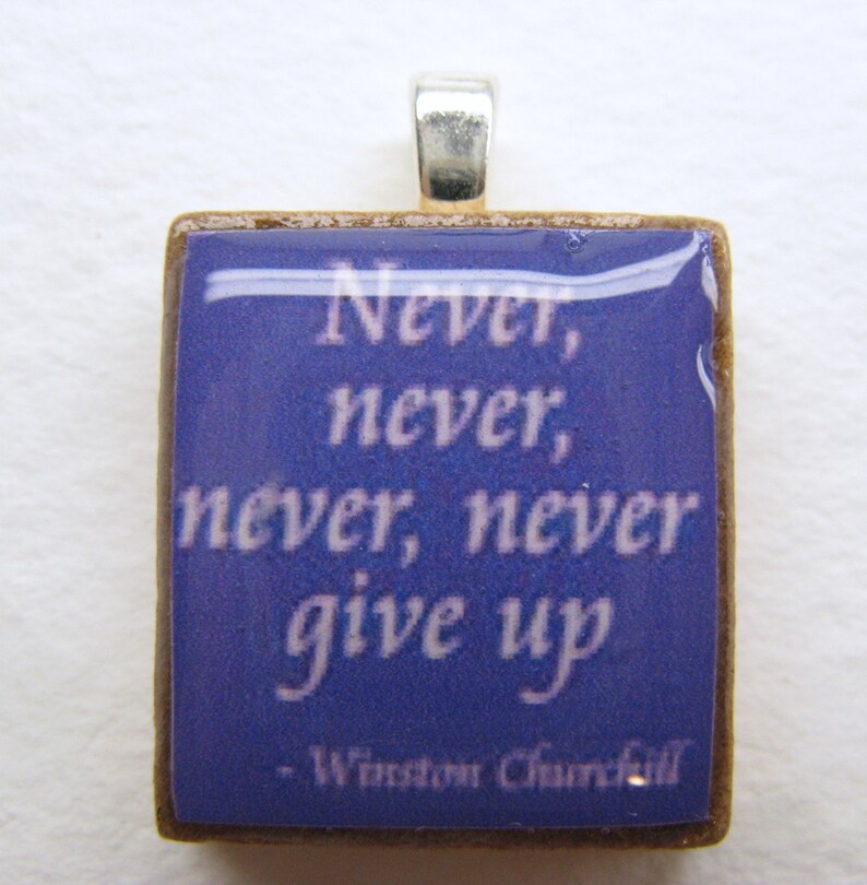 Puede incluir: Un colgante cuadrado con fondo azul y texto blanco que dice "Never, never, never, never give up - Winston Churchill". El colgante est&aacute; hecho de madera y tiene una anilla de plata.