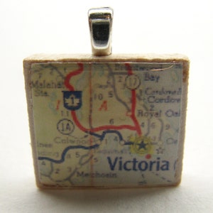 May include: A square, cream-colored pendant with a vintage map of Victoria, Texas. The map features a red highway, blue roads, and a star marking the location of Victoria. The pendant has a silver bail.