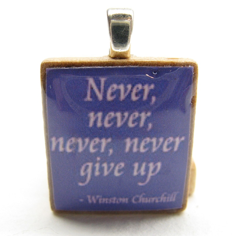 Puede incluir: Un peque&ntilde;o colgante cuadrado con una anilla de plata. El colgante est&aacute; hecho de un material marr&oacute;n claro y tiene un fondo azul con una cita blanca que dice "Never, never, never, never give up - Winston Churchill".