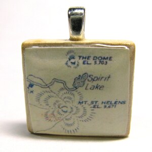 May include: Square pendant with a map design featuring Spirit Lake and Mt. St. Helens, with the text "THE DOME EL. 5.703" and "MT. ST. HELENS EL. 9.671". The pendant has a silver bail and a light-colored wooden frame.