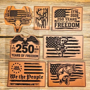 May include: A collection of six tan leather patches featuring black laser-engraved designs. The patches display American flag motifs, deer skulls, a bald eagle, a fish, and text including 1776-2026, 250 Years of Freedom, and We the People.