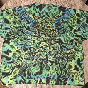 May include: A tie-dye t-shirt with a vibrant pattern of green, black, and blue hues. The design features a repeating leaf-like motif, creating a visually dynamic and textured appearance. The shirt is laid flat, showcasing its round neck and short sleeves.