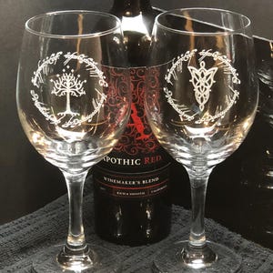 Puede incluir: Dos copas de vino transparentes con diseños grabados del Anillo Único de El Señor de los Anillos. La copa de la izquierda presenta el Árbol Blanco de Gondor, mientras que la copa de la derecha presenta el símbolo élfico del Anillo Único. Una botella de vino tinto está en el fondo.