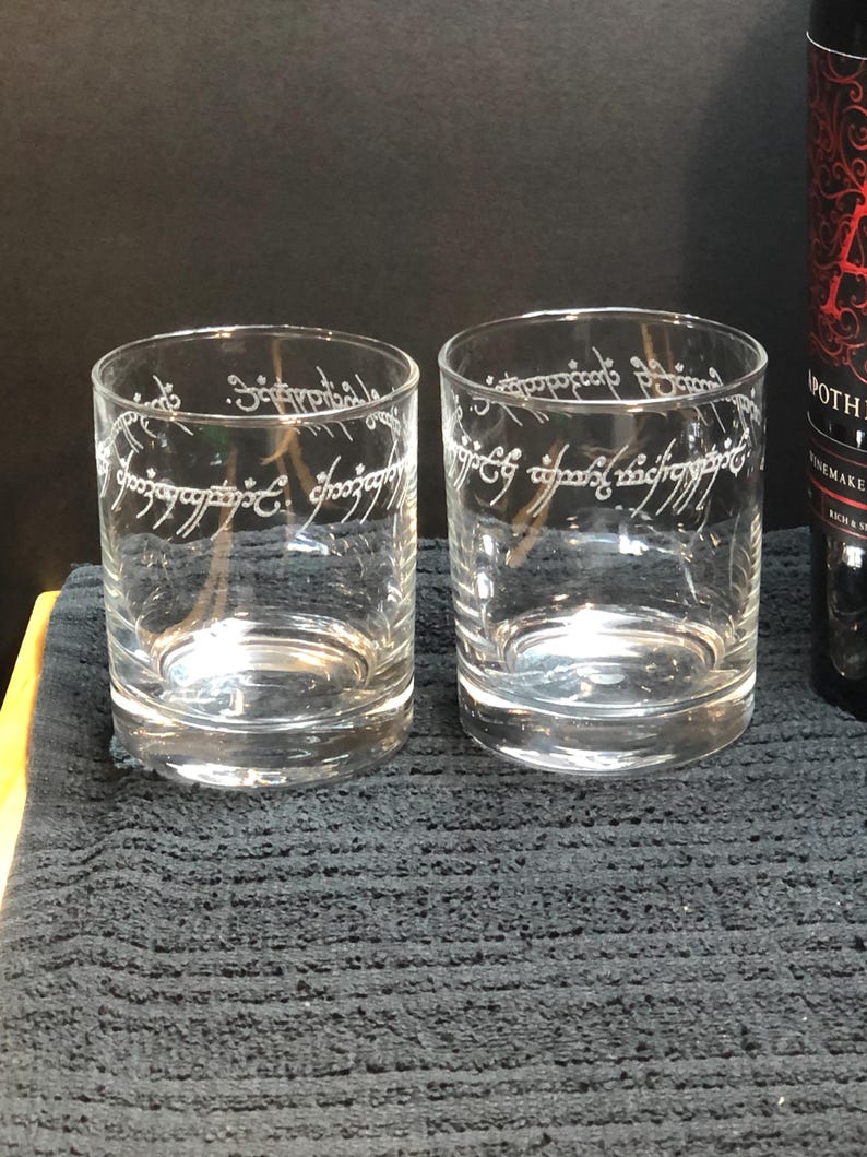 May include: Two clear glass tumblers with the inscription "One Ring to rule them all" etched on the side. The inscription is in the Elvish language from the Lord of the Rings.