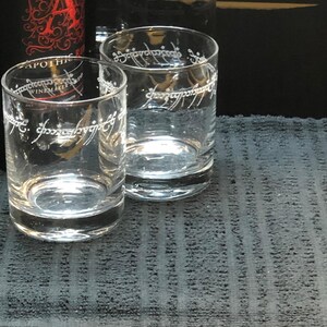 May include: Two clear glass tumblers with the inscription "One Ring to rule them all" etched on the side. The glasses are sitting on a dark gray textured surface.