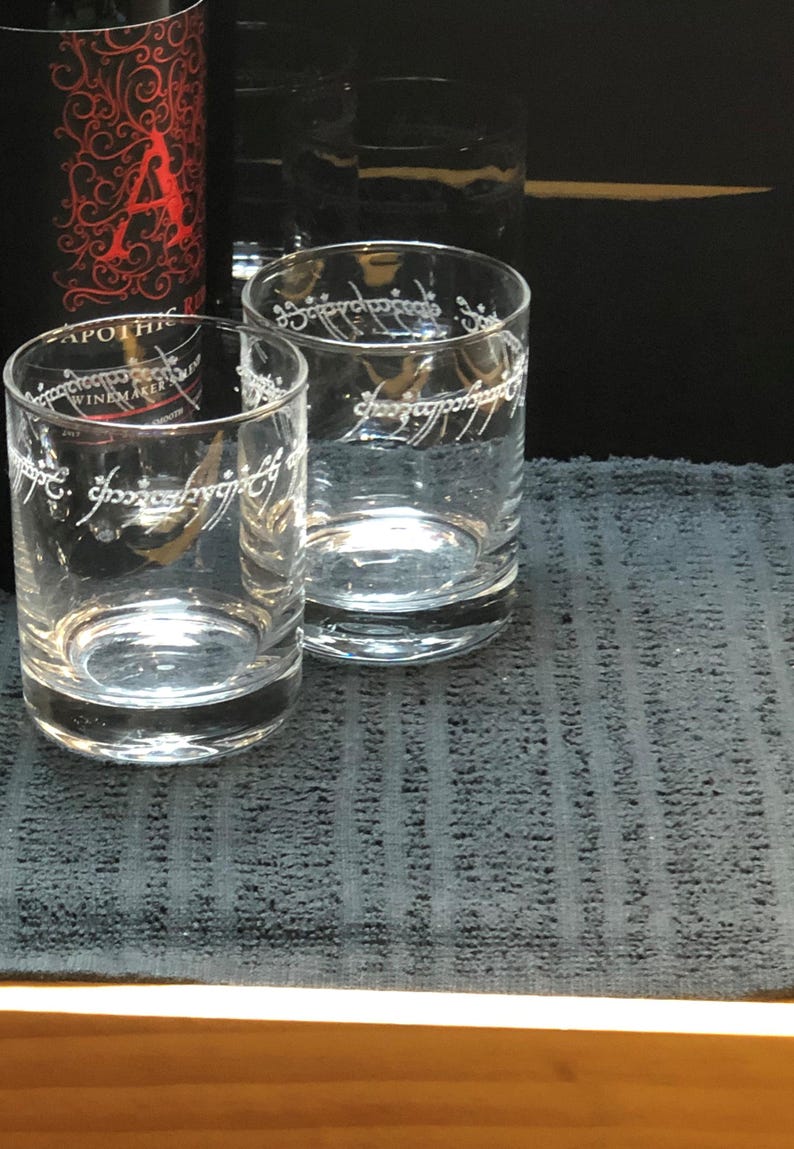May include: Two clear glass tumblers with the inscription "One Ring to rule them all" etched on the side. The glasses are sitting on a dark gray textured surface.