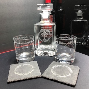 May include: A clear glass decanter with a stopper, two matching glasses, and two square coasters. The decanter and glasses feature etched designs. The coasters are dark gray with a white etched design. The theme appears to be related to a fantasy series.