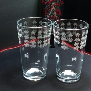 May include: Two clear glass pint glasses with an etched design of white space invaders. The glasses are on a black surface.