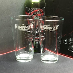 Könnte beinhalten: Zwei transparente Biergläser mit dem Text "THE BRONZE" in weißer Schrift und zusätzlichem Text. Hinter den Gläsern ist eine grüne Weinflasche zu sehen. Die Gläser stehen auf einer dunklen Oberfläche.