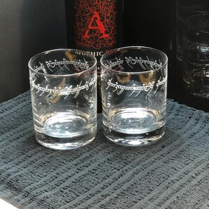 May include: Two clear glass tumblers with the inscription "One Ring to rule them all" etched on the side. The glasses are sitting on a black textured surface.