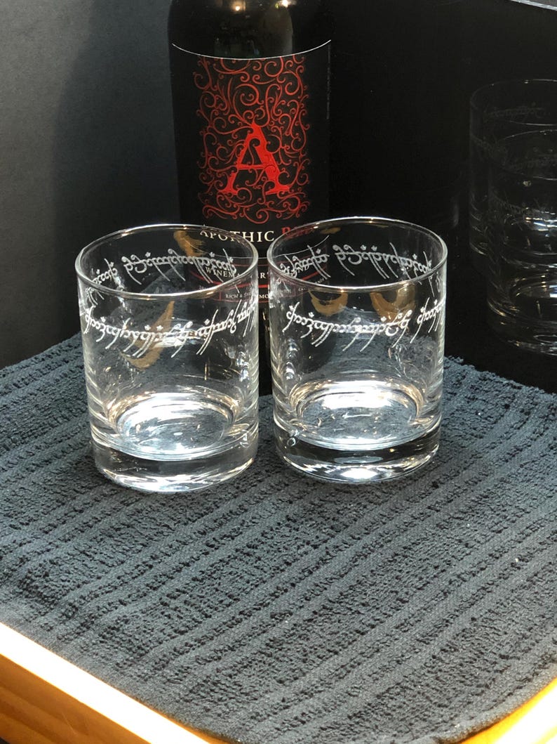 May include: Two clear glass tumblers with the inscription "One Ring to rule them all" etched on the side. The glasses are sitting on a black textured surface.