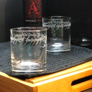 May include: Two clear glass tumblers with the inscription "One Ring to rule them all" etched on the side. The glasses are sitting on a wooden tray with a black cloth napkin.