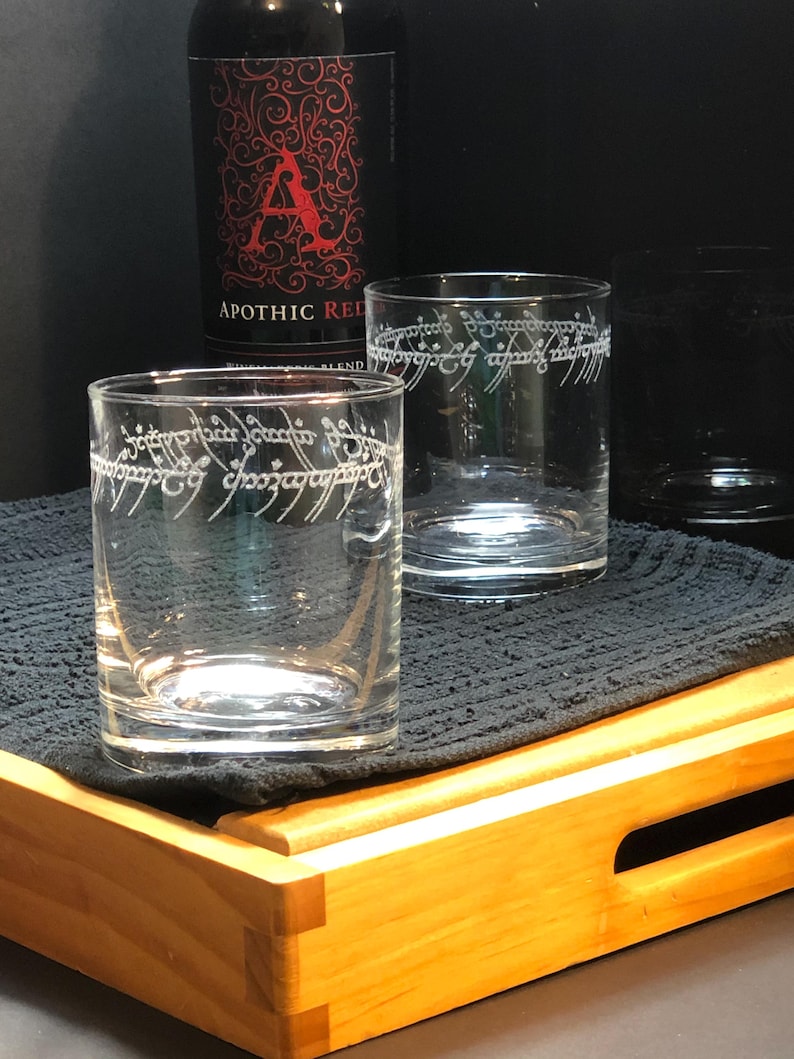 May include: Two clear glass tumblers with the inscription "One Ring to rule them all" etched on the side. The glasses are sitting on a wooden tray with a black cloth napkin.