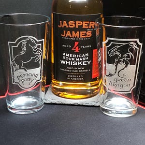 May include: Three glasses with etched designs: a prancing pony, a green dragon, and a bottle of Jasper James 4 year aged American Sour Mash Whiskey.