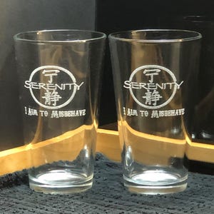 Könnte beinhalten: Zwei klare Pintgläser aus Glas mit einem weißen, geätzten Design. Das Design beinhaltet das Wort "Serenity" in einem Kreis mit einem chinesischen Schriftzeichen in der Mitte. Der Text "I aim to misbehave" befindet sich unterhalb des Kreises.