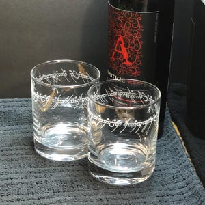 May include: Two clear glass tumblers with the inscription "One Ring to rule them all" etched around the top. The glasses are sitting on a black textured surface next to a bottle of red wine.