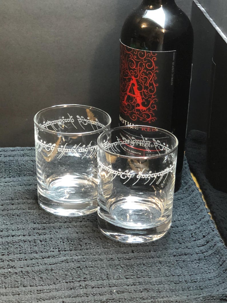 May include: Two clear glass tumblers with the inscription "One Ring to rule them all" etched around the top. The glasses are sitting on a black textured surface next to a bottle of red wine.