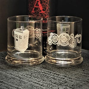 May include: Two clear glass tumblers with a white design. One tumbler features a white silhouette of a police box, the other features a white design of concentric circles. The tumblers are sitting on a black textured surface.