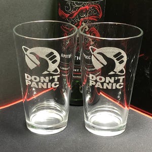 May include: Two clear glass pint glasses with the words "Don't Panic" etched on the front. Each glass features a stylized image of a planet with a hand giving a thumbs up.