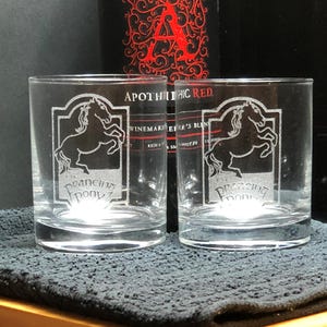 Può includere: Due bicchieri di vetro trasparente con immagini incise di un pony al galoppo. I bicchieri sono su una superficie nera. Il testo sui bicchieri recita "Prancing Pony".