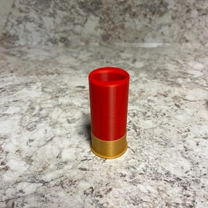 May include: A red and gold shotgun shell casing. The casing is cylindrical with a red body and a gold-colored base. The object is standing upright on a speckled surface.