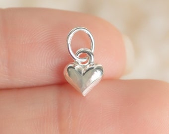 May include: A small silver charm in the shape of a heart. The heart is slightly puffed out and has a smooth, shiny finish.
