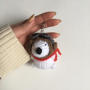 May include: White crocheted plush toy wearing a brown aviator hat with a red scarf. The toy is attached to a silver keychain.