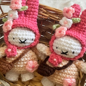 May include: Two handmade crochet bunny dolls in a woven basket. The bunnies have white faces, pink hooded hats adorned with pink and white flowers, and small brown satchels. The bunnies' bodies are beige and white.