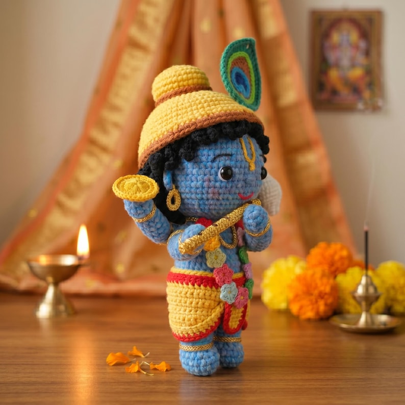 Puede incluir: Mu&ntilde;eca de ganchillo azul de una deidad, que lleva un sombrero y prendas amarillas con detalles dorados. La mu&ntilde;eca sostiene una flauta y un objeto circular. Una pluma de pavo real est&aacute; unida al sombrero. El fondo incluye una vela, flores y una imagen enmarcada.