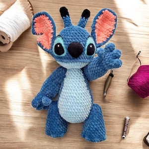 Könnte beinhalten: Blaues gehäkeltes Plüschtier mit großen Ohren, einer schwarzen Nase und großen Augen. Das Spielzeug hat die Form eines Comic-Charakters mit einem blauen und hellblauen Körper.