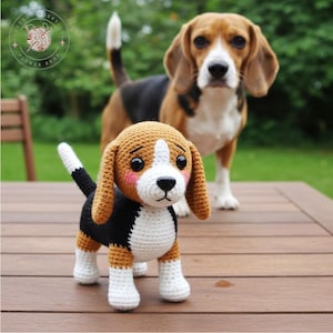 Könnte beinhalten: Gehäkelter Beagle-Hund mit braunen, schwarzen und weißen Details, der auf einer Holzoberfläche steht. Ein echter Beagle-Hund steht im Hintergrund. Der gehäkelte Hund hat eine weiße Schnauze, rosa Wangen und Schlappohren.