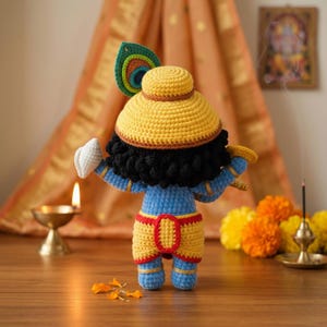 Puede incluir: Mu&ntilde;eca de ganchillo hecha a mano de una deidad, con sombrero amarillo, piel azul y ropa amarilla y roja. La mu&ntilde;eca tiene pelo negro y un detalle de pluma de pavo real. Se muestra con una flauta y un gesto de mano blanca.
