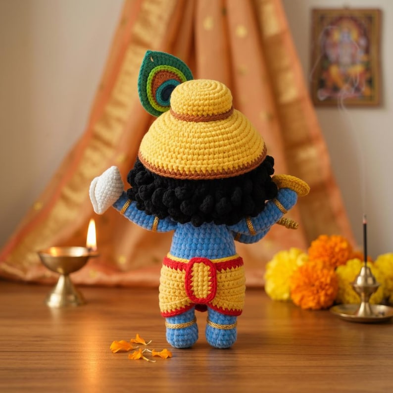 Puede incluir: Mu&ntilde;eca de ganchillo hecha a mano de una deidad, con sombrero amarillo, piel azul y ropa amarilla y roja. La mu&ntilde;eca tiene pelo negro y un detalle de pluma de pavo real. Se muestra con una flauta y un gesto de mano blanca.