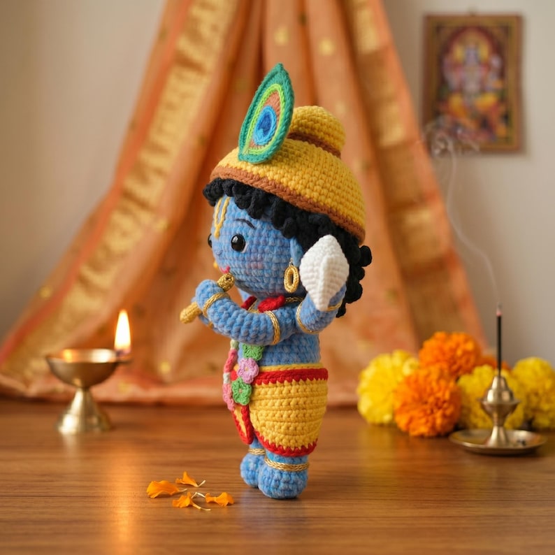 Puede incluir: Mu&ntilde;eca de Krishna hecha a mano con ganchillo, con cuerpo azul, ropa amarilla y una pluma de pavo real en su sombrero amarillo. La mu&ntilde;eca sostiene una flauta y un objeto blanco, con una guirnalda alrededor del cuello. El fondo incluye una vela, flores y una cortina decorativa.