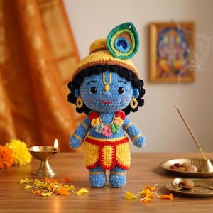 Puede incluir: Mu&ntilde;eca de ganchillo azul de Krishna, con un sombrero amarillo con una pluma de pavo real, una guirnalda floral y ropa amarilla y roja. La mu&ntilde;eca tiene pelo negro y pendientes. Una peque&ntilde;a l&aacute;mpara de aceite e incienso est&aacute;n en el fondo.