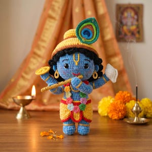 Puede incluir: Mu&ntilde;eca de Krishna azul tejida a ganchillo, con turbante amarillo y pluma de pavo real. La mu&ntilde;eca sostiene una flauta y una caracola. Est&aacute; adornada con detalles dorados y una guirnalda floral. Fondo con l&aacute;mpara encendida y flores.