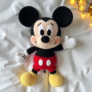 Può includere: Un peluche di Topolino fatto a mano all'uncinetto. Il giocattolo presenta orecchie, testa e braccia nere, un viso color crema e un abito rosso e bianco. Il peluche è appoggiato su una superficie bianca.
