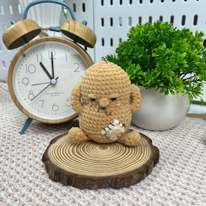 May include: A handmade, beige crochet doll with a grumpy expression, small arms, and a stitched mouth. The doll sits on a small wooden slice. A vintage-style alarm clock and a small potted plant are in the background.