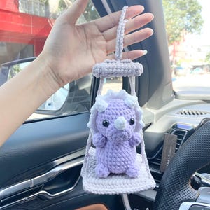May include: A handmade, lavender-colored crochet Triceratops dinosaur car charm. The dinosaur is suspended in a light gray crochet swing, with a white frill around the head. The charm is designed to hang from a car's rearview mirror.