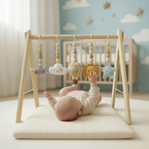 May include: A wooden baby gym featuring hanging crochet toys: a plane, cloud, hot air balloon, sun, and star in pastel shades. A baby lies on a cream mat, reaching for the yellow sun. The gym is set up in a nursery.