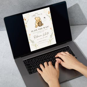 May include: A laptop screen showing a baby shower invitation with a Winnie the Pooh design. The invitation says "Our little honey, IS ON THE WAY!" with the name "Rebecca White" and event details. Hands are typing on the laptop keyboard.
