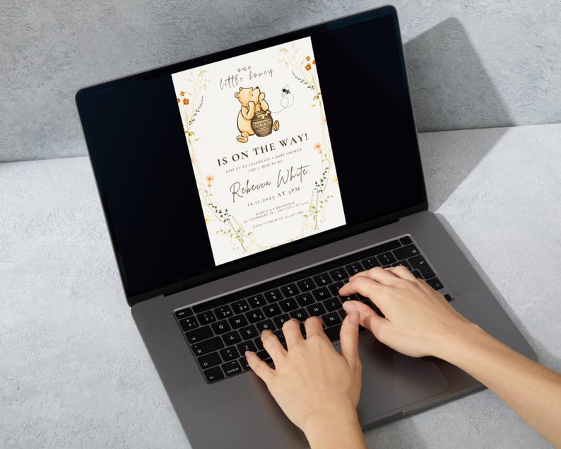 May include: A laptop screen showing a baby shower invitation with a Winnie the Pooh design. The invitation says "Our little honey, IS ON THE WAY!" with the name "Rebecca White" and event details. Hands are typing on the laptop keyboard.