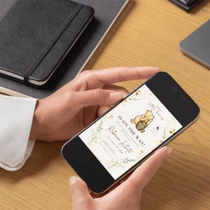 May include: A smartphone screen shows a baby shower invitation with a Winnie the Pooh theme. The invitation reads "Is on the Way!" and includes the name "Rebecca White". The design incorporates floral elements and a honey pot.