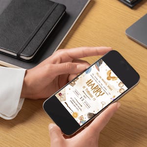 May include: A smartphone displaying a birthday invitation with the text "Harry is turning 13." The invitation features illustrations of a golden snitch, a key, and a broom. The phone is held by a person, with a notebook and laptop in the background.