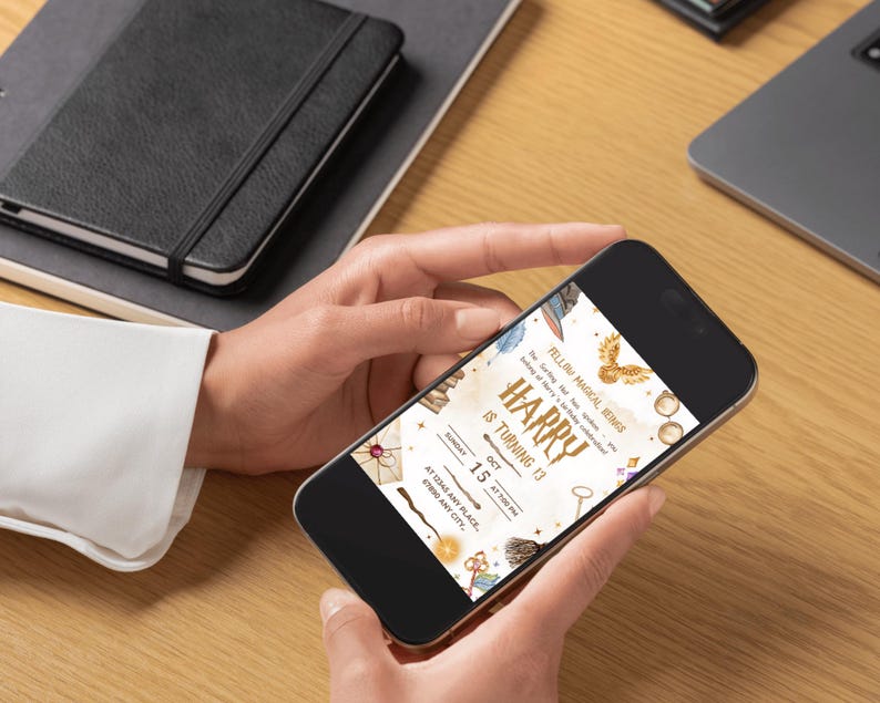 May include: A smartphone displaying a birthday invitation with the text "Harry is turning 13." The invitation features illustrations of a golden snitch, a key, and a broom. The phone is held by a person, with a notebook and laptop in the background.
