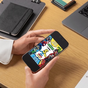 May include: A smartphone displaying a colorful birthday invitation with superhero graphics. The invitation features the text "Connor is turning 5" in a comic book style. The phone is held by a person on a wooden desk, with a laptop and notebook in the background.