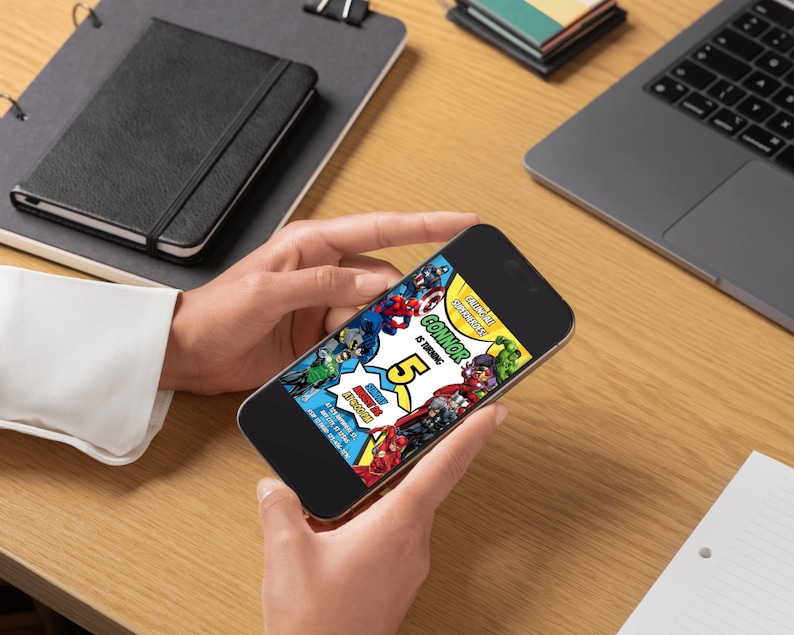May include: A smartphone displaying a colorful birthday invitation with superhero graphics. The invitation features the text "Connor is turning 5" in a comic book style. The phone is held by a person on a wooden desk, with a laptop and notebook in the background.