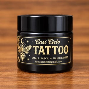 Puede incluir: Un tarro cilíndrico negro con tapa de rosca negra. La etiqueta presenta texto dorado que dice Casi Cielo TATTOO, SMALL BATCH, HANDCRAFTED y hey.casicielo@gmail.com, acompañado de un gráfico dorado de una abeja, una luna creciente y estrellas.