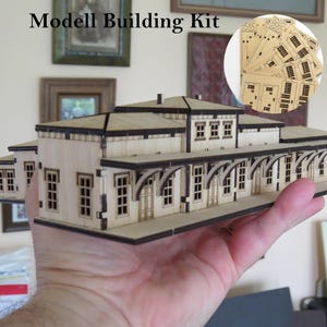 May include: A laser-cut wooden model building kit of a train station featuring a central structure with a roof, multiple windows, and a platform with arched supports. An inset circle shows several flat, un-assembled wooden sheets with cut-out parts.