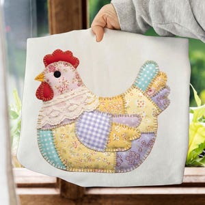 Op de afbeelding: Een decoratief patchwork kippenontwerp op een witte achtergrond. De kip heeft een rode kam en lellen, een gele snavel en een bloemmotief op de kop. Het lichaam bestaat uit verschillende stoffen lappen in geel, paars en blauw, met een kanten kraag.